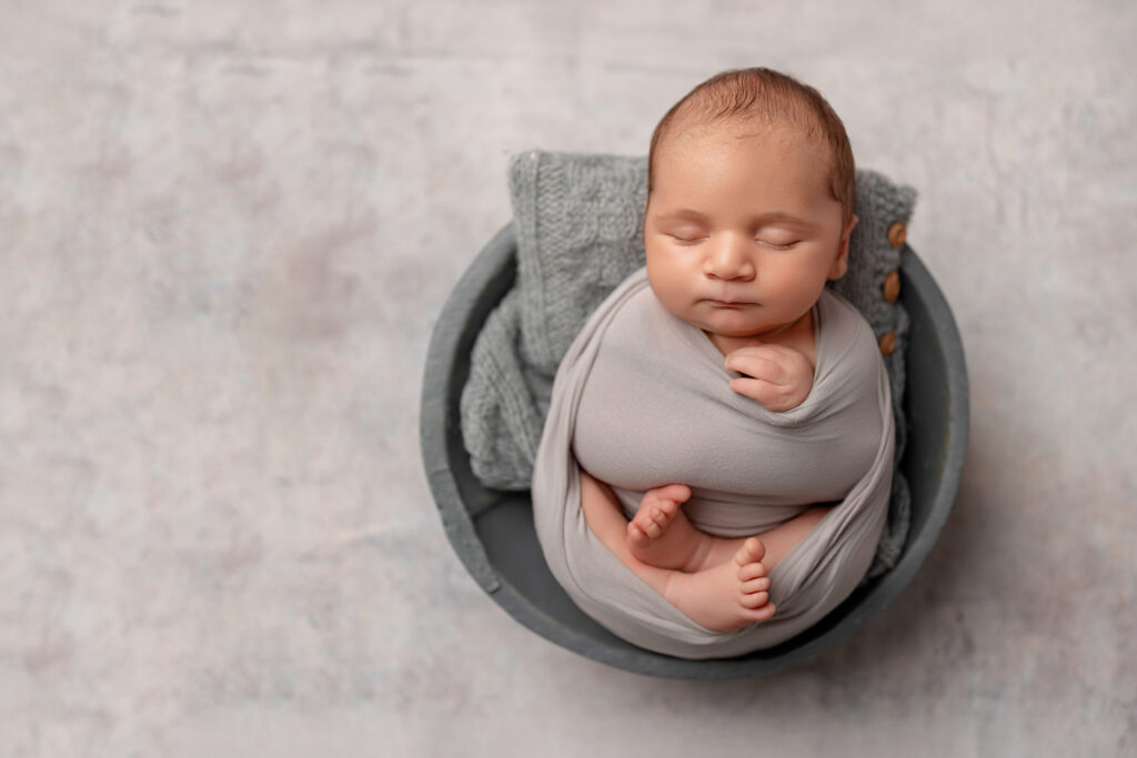 Wrapped newborn baby boy posed in a grey bowl with soft grey layers in a top-down portrait.