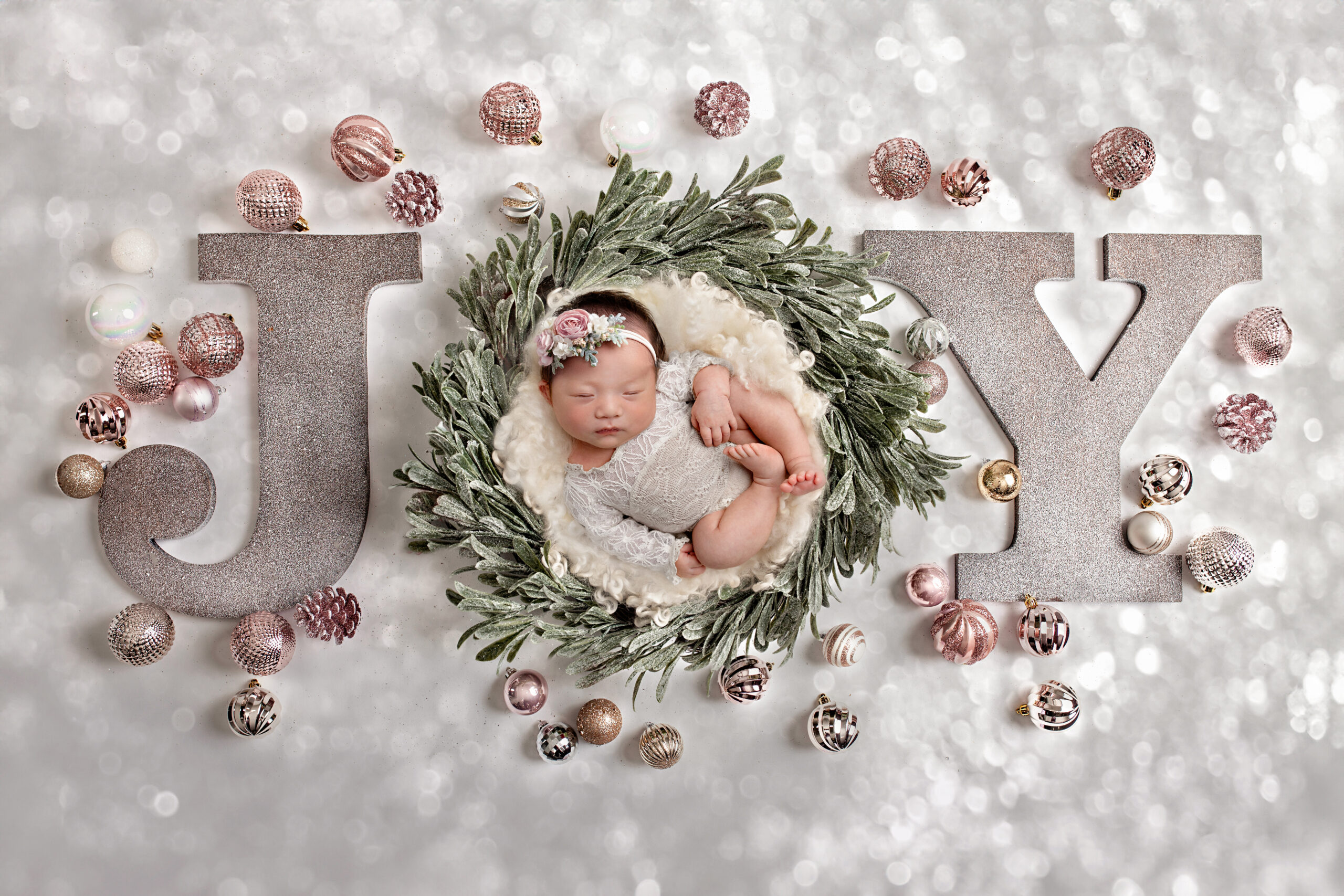 Newborn sleeping in a greenery wreath surrounded by the word “JOY,” styled in soft neutral Christmas tones during a timeless studio newborn session.