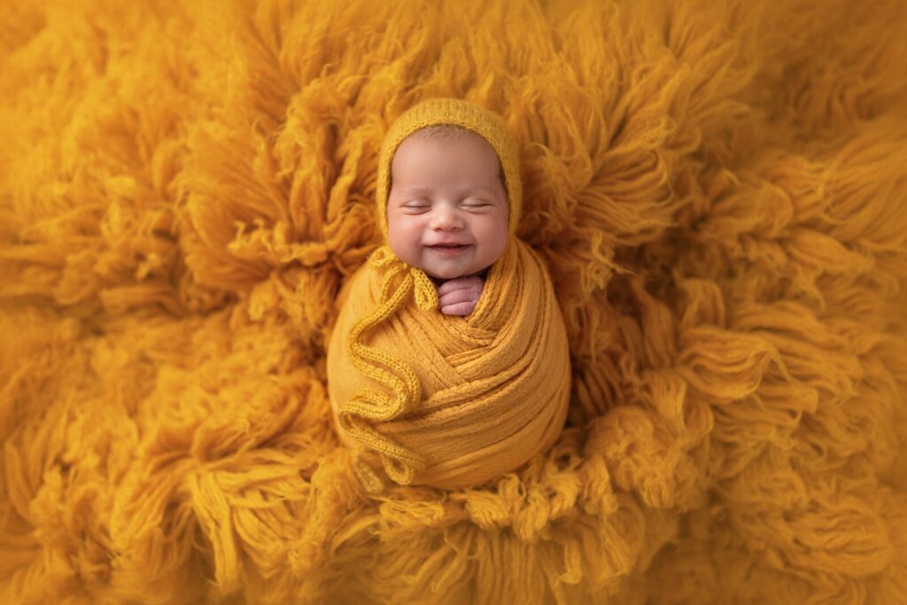 Infant smiles in sleep decked out in a deep gold wrap and surrounded by a fluffy gold background at an indoor photography location in Mississauga​