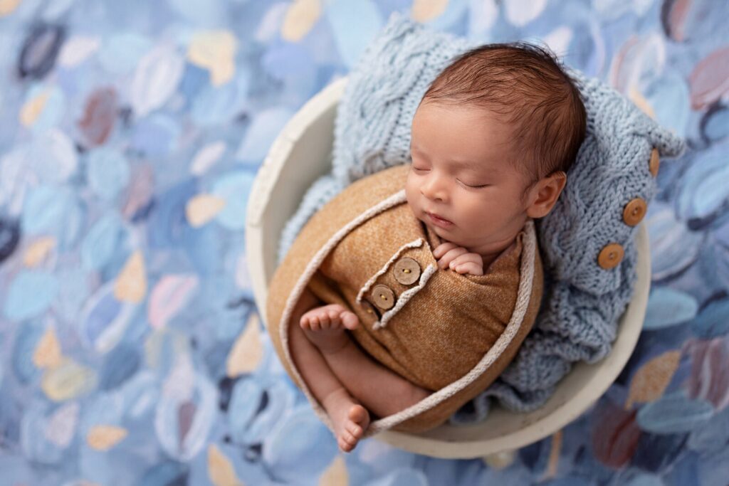 One of the best photography studios in Mississauga showcases baby boy sleeping in woven basket with blue and yellow textured background. 