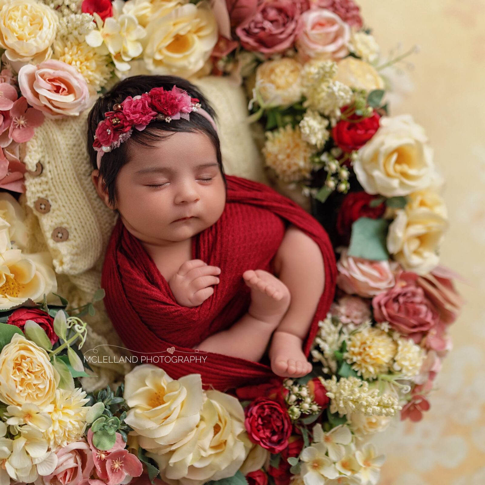 Professional newborn portrait of a sleeping baby wrapped in red and surrounded by flowers