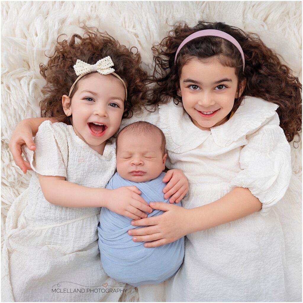 Two older sisters cuddling their sleeping newborn sibling on a cream blanket