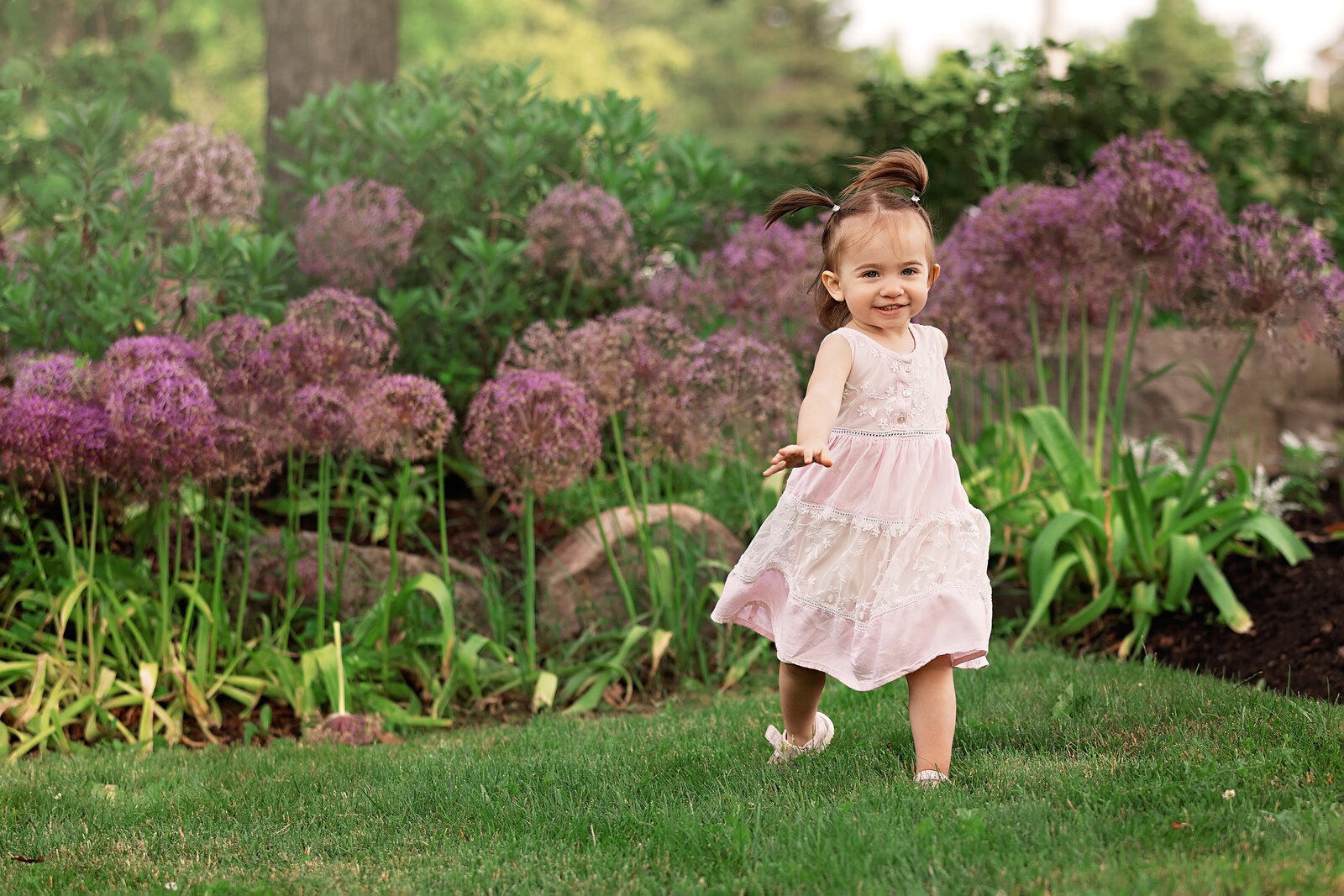 Smiling toddler in a pale pink dress walking across the grass in front of purple flowers during an outdoor photo session