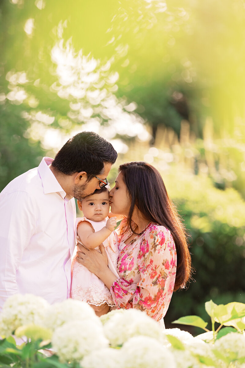 Parents kissing their young daughter on both cheeks while holding her beneath a tree during an outdoor family photography session